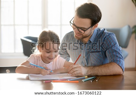 Head shot joyful young dad drawing pictures in paper album with happy small daughter, enjoying free leisure time at home. Smiling little kid girl learning painting with father, sitting at table.