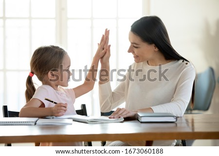 Joyful small daughter giving high five to satisfied with homework results mother. Happy young mom teacher babysitter praising little preschool child girl for making right tasks, sitting at home. Royalty-Free Stock Photo #1673060188