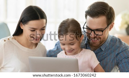 Head shot close up happy married couple relaxing with little daughter, using computer together at home. Smiling parents watching funny videos cartoons with kid girl, shopping making purchases online.