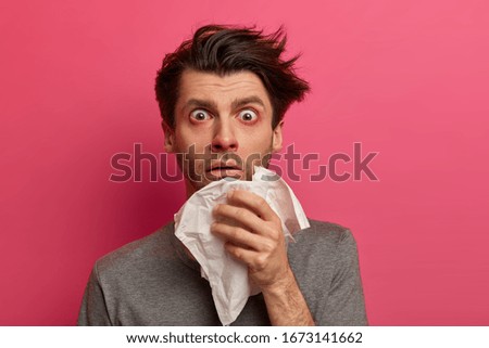 Stunned sick man has flu, virus or allergy respiratory, red watery eyes, blows nose in tissue, finds out about serious disease, poses over pink background. Health, medicine and symptoms concept Royalty-Free Stock Photo #1673141662