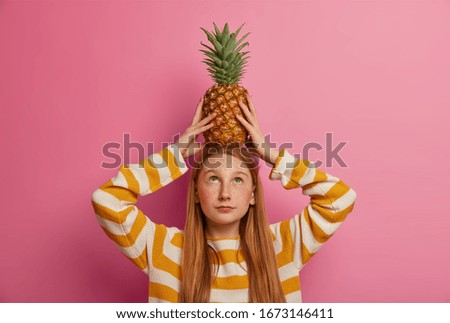 Little pretty female child holds ripe pineapple on head, has long red hair, dressed in striped jumper, poses indoor against pink background. Adorable freckled kid with tropical fruit, makes trick