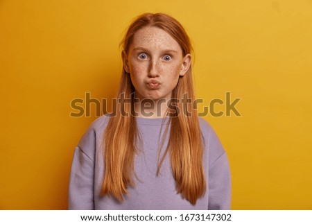 Attractive shocked girl keeps lips rounded, has surprised grimace, gazes directly at camera, wears casual jumper, has big ears, freckled skin, isolated on yellow background. Face expressions