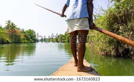 A picture of a brown skin tone man in a bright blue shirt and beige pants rowing a wooden boat with a bamboo paddle surrounded by a beautiful nature scenery of backwater and lush green trees shore.