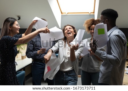Happy excited diverse employees dancing with papers documents in hands, successful team having fun after contract signing, celebrating business success, deal, achievement at meeting in modern office