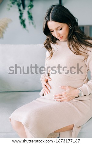 portrait of a young beautiful pregnant woman in a beige dress sitting on a gray sofa in a cozy Studio