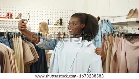 Stylish young African American woman in store and putting to herself clothes on hangers instead of trying on. Pretty female buyer shopping in fashion shop taking selfie photo with phone camera.