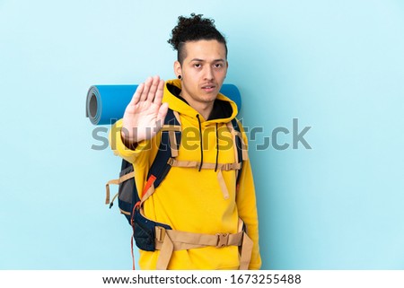 Young mountaineer man with a big backpack over isolated blue background making stop gesture with her hand