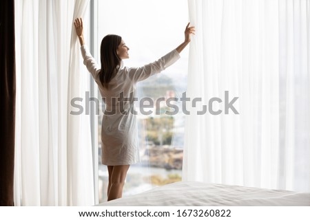 Attractive energetic happy young brunette woman wearing nightgown bathrobe, opening curtains on big panoramic window. Smiling lady enjoying starting of sunny day holiday vacation morning time. Royalty-Free Stock Photo #1673260822