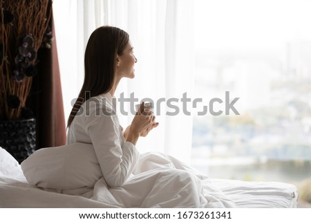 Side view happy attractive young brunette woman sitting in bed under duvet, holding cup of black coffee, enjoying peaceful calm weekend vacation morning time alone in bedroom at home or hotel.