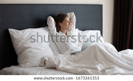 Happy pretty young woman crossed hands behind head, enjoying peaceful good morning time under duvet with computer. Smiling lady in pajamas sitting in cozy bed, planning day, dreaming of future.