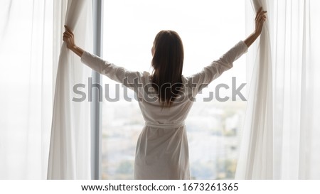 Back rear view confident woman in night robe gown pulling opening lace curtains after waking up in morning, admiring panorama view near big window, enjoying start of new day at home or hotel. Royalty-Free Stock Photo #1673261365