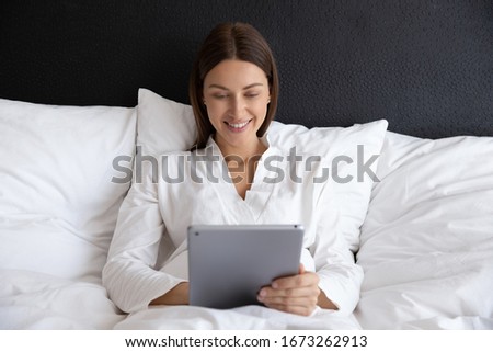 Front view attractive smiling young woman lying on pillows in comfortable bed, using digital tablet after waking up, reading electronic book, enjoying spending weekend vacation good morning time.