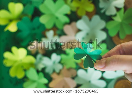 Womans hand holds green four-leafed paper shamrock on blurred background with different shades of green four-leafed paper clovers with chocolate coins arranged in chaotic order. Lucky concept