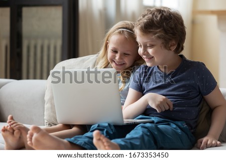 Funny smart small siblings sit on comfortable couch at home using laptop watching cartoon at home together, clever little kids brother and sister have fun enjoy movie or video on computer