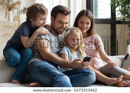 Happy young family with little children sit relax on comfortable couch watching funny video on smartphone together, smiling parents with kids rest on sofa at home enjoying cartoon on cellphone