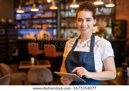 Young smiling waitress in apron and t-shirt standing in front of camera while using touchpad and meeting guests in cafe Royalty-Free Stock Photo #1673354077