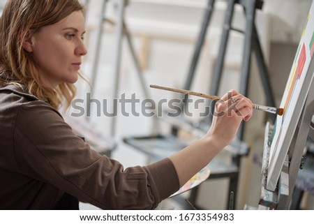 Young serious woman with paintbrush looking at unfinished picture while sitting in front of easel and painting at lesson