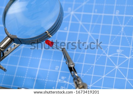 Close-up of red led electrical component held by crocodile-clips and seen through magnifying glass on blue background. Concept of electronics, science, industry, hobby or education.