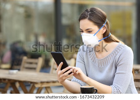Serious woman with protective face mask looking at smart phone checking news on a cafe terrace