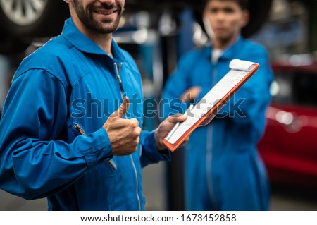 Close up thumbs up hand sign of professional look technician inspecting car underbody and suspension system by using check list in modern car service shop. Automotive business or car repair concept.