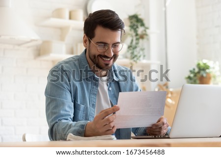 Smiling young man reading good news in paper notification, sitting at table with laptop, happy satisfied male wearing glasses holding document, job promotion, loan approval, great exam result Royalty-Free Stock Photo #1673456488