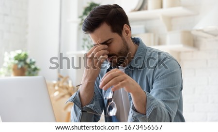 Tired unhealthy man taking of glasses, massaging nose bridge, feeling unwell, suffering from eye strain after long laptop use, exhausted young male student, freelancer relieving dry eyes syndrome Royalty-Free Stock Photo #1673456557