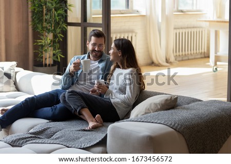 Loving couple relaxing on cozy sofa in living room, drinking hot beverages, tea or coffee, happy smiling man and woman holding white cups sitting on couch, chatting, talking, spending lazy weekend Royalty-Free Stock Photo #1673456572