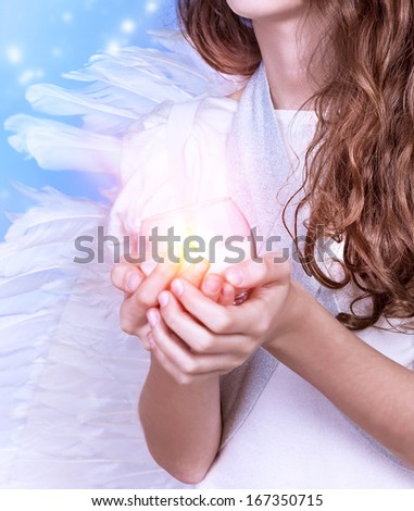 Closeup on little angel holding in hands candle, body part of teen girl wearing white dress and big fluffy wings, Christmas celebration, religious holiday concept