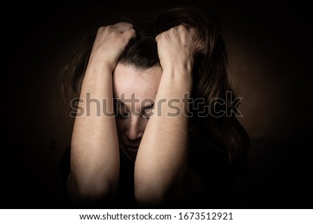 Low key photo of depressed young crying woman, victim of domestic violence. Scared girl sitting with head in her hands. Emotions in female face. Violence against women.

