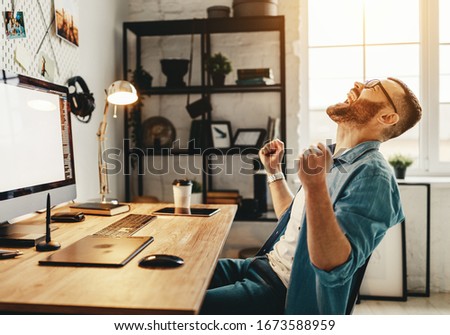 Side view of overjoyed young male in casual clothes triumphing success with raised fists while sitting in front of large monitor at workplace
