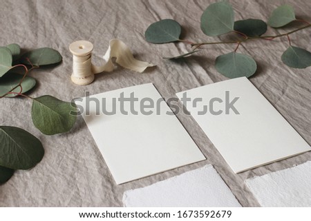 Wedding stationery mock-up scene. Blank greeting cards on linen tablecloth background with green eucalyptus tree leaves and branches and silk ribbon. Feminine still life composition. High angle view.