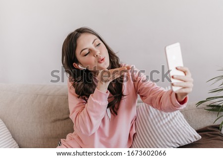 Woman in sweatshirt and wireless headphones blows kiss and takes selfie. Portrait of brunette curly teen girl in pink outfit talking by video call at home