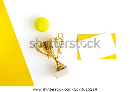 Champion trophy cup, tennis ball, envelope and blank sheet for text on a geometric white and yellow background. Winning concept. Top view. Flat lay. Sports layout and tabletop mockup with copy space.