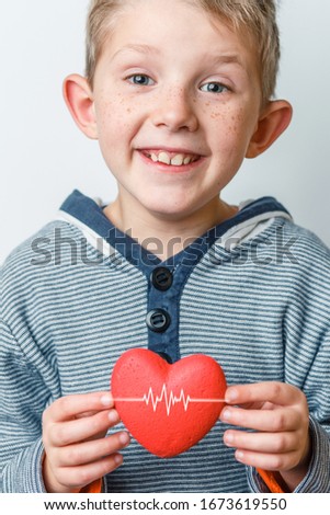 world health day. Smiling child with freckles holds a red heart with both hands. In a striped sweater on a gray background. Studio photography.