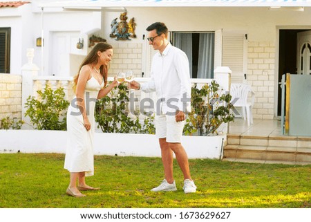 A couple drinks white wine on the lawn near their home on the island. On a trip, the couple rented a house, relax in the sun, the couple laughs and drinks wine. A young couple celebrates a purchase at
