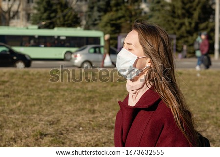 Girl Portrait of a girl in a red coat. She closed her eyes from a headache. On the face is a medical protective mask. Protection against bacteria, viruses. Symptom of coronavirus. Street Royalty-Free Stock Photo #1673632555
