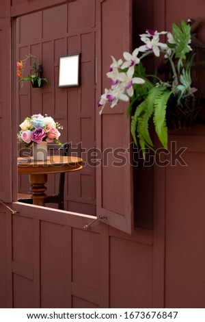 Side view and selective focus at artificial rose flowers in ceramic vase on wooden round table with blank picture frame on wooden wall in window frame and blurred foreground, home decoration concept