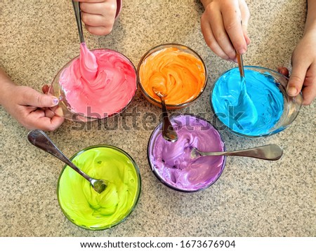 Two Little Girls' Hands Mixing Bright, Colorful Frosting in Small Glass Bowls with Small Metal Spoons - Pink, Blue, Purple, Green, and Orange; Kids Baking