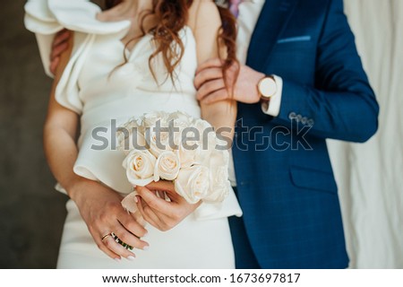 Wedding bouquet of fresh white roses in bride hands. Stylish wedding picture. groom in a blue suit gently embraces the bride in a white stylish dress made of stiff fabric with a Puffy flounce sleeve.