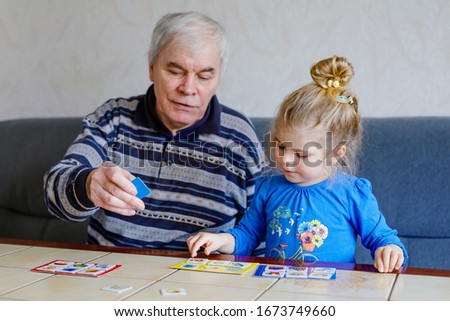 Beautiful toddler girl and grandfather playing together pictures lotto table cards game at home. Cute child and senior man having fun together. Happy family indoors