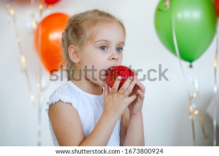 Girl blue-eyed blonde eats a red apple. Against the background of festive multi-colored balloons
