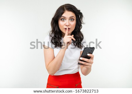 girl keeps a secret while holding a finger at her mouth and a smartphone in her hand on a white background