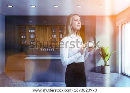 Thoughtful blonde businesswoman with coffee standing in spacious kitchen with grey walls, concrete floor, wooden cupboards, gray countertops and comfortable island. Toned image
