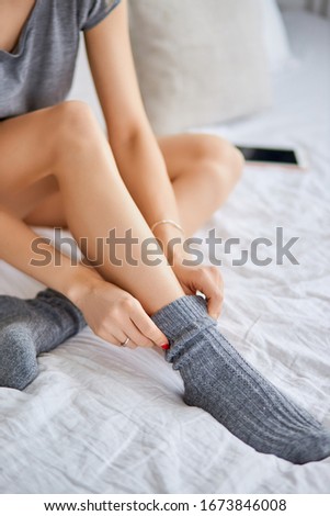 cropped photo of young lady sitting on bed in light bedroom, lady with slender legs neatly puts on gray socks.