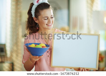 Smiling modern woman in a pink blouse and easter bunny ears with a blue plate with colorful easter eggs with a blue plate with colorful easter eggs in the modern living room in sunny spring day.