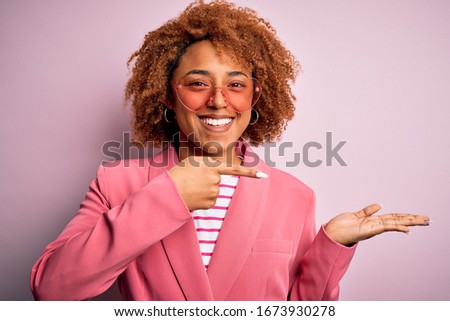 Young African American afro woman with curly hair wearing funny hearts sunglasses amazed and smiling to the camera while presenting with hand and pointing with finger.
