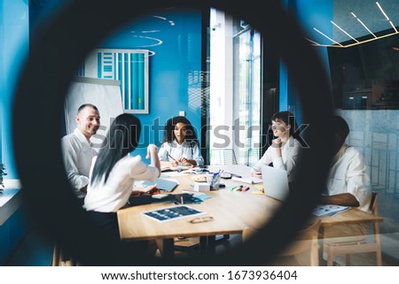 Through glass view of smiling multiracial coworkers in white shirts talking and gesturing while sitting at conference table with charts and papers beside flip chart at urban office