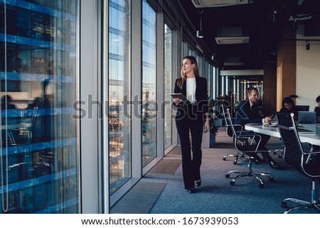 Serious female administrative manager in formal suit walking in office with digital tablet for checking office working process, confident woman business owner looking at window passing with touchpad Royalty-Free Stock Photo #1673939053