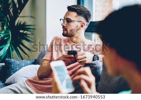 Young caucasian hipster guy in eyewear looking away using mobile phone for chatting sitting next to girlfriend at home, cropped image with blurred frontage of woman millennial holding smartphone