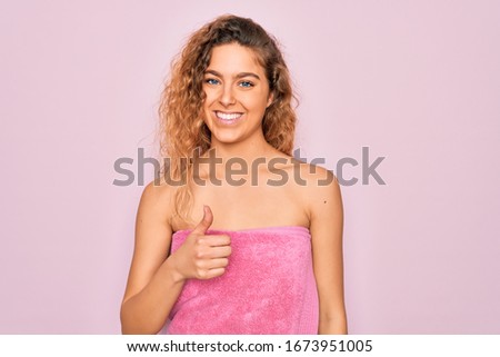 Beautiful blonde woman with blue eyes wearing towel shower after bath over pink background doing happy thumbs up gesture with hand. Approving expression looking at the camera showing success.